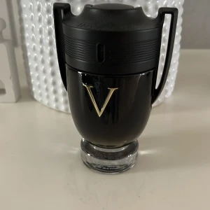 Svart parfymflaska med guld V - Snygg och stilren svart parfymflaska med ett stort guld V på framsidan. Flaskan har en unik design som påminner om en pokal med två handtag på sidorna och en svart kork. Perfekt för den som vill ha en elegant och maskulin doft på hyllan.