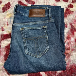 Blå jeans från Tiger of Sweden - Snygga blå jeans från Tiger of Sweden med slitna detaljer och en klassisk femficksdesign. I storlek 29/30. De har en läderpatch med logotyp baktill och en dragkedja framtill. Perfekta för en avslappnad stil. Ett litet hål i högerfickan 