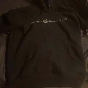 Svart hoodie från Sail Racing - Säljer en svart hoodie från Sail Racing med logga på bröstet. Tröjan har en klassisk design med huva och fickor framtill. Perfekt för en avslappnad stil.