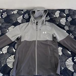 Grå vindjacka från Under Armour - Snygg grå vindjacka från Under Armour med huva och dragkedja framtill. Jackan har långa ärmar och elastiska muddar med logotypdetaljer. Perfekt för blåsiga dagar.