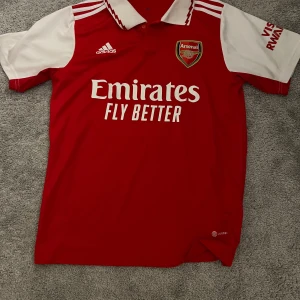Röd Arsenal fotbollströja från Adidas - Säljer en snygg röd Arsenal fotbollströja från Adidas med vita detaljer och klubbens emblem på bröstet. Tröjan har korta ärmar och en klassisk krage. Perfekt för alla Arsenal-fans! 🔴⚪