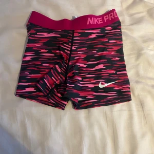  Nike PRO Shorts - Säljer ett par färgglada träningsshorts från Nike Pro. De har ett mönster i rosa och svart med en vit Nike-logga. Perfekta för träning med en tight passform och elastisk midja.