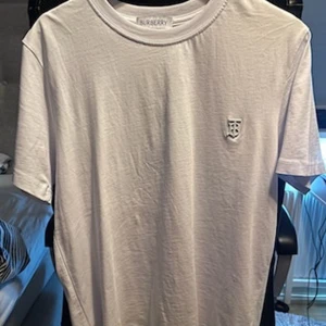 Vit t-shirt från Burberry med metall logga - Säljer en stilren vit t-shirt från Burberry med en broderad logotyp på bröstet. T-shirten har en klassisk rund hals och korta ärmar. Perfekt för en minimalistisk look.