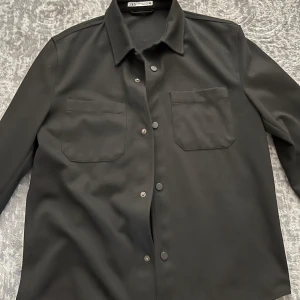 Svart overshirt från Zara - Snygg svart overshirt från Zara med klassisk krage och knappar framtill. Två bröstfickor ger en stilren look. Perfekt för lager-på-lager-stil.