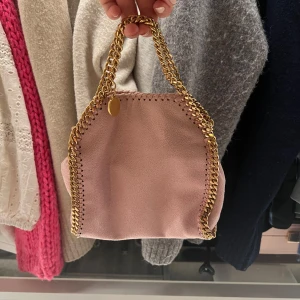 Rosa axelväska från Stella McCartney - Säljer en snygg rosa axelväska från Stella McCartney med guldfärgade kedjedetaljer. Väskan har en elegant design och är perfekt för att lyfta din outfit. Den är tillverkad i syntetmaterial och har en praktisk storlek för dina nödvändigheter. Inga som helst tecken på användning! Pris kan diskuteras