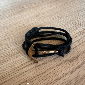 Svart armband med guldfärgad ankare - Snyggt svart armband från Blvckout med flera varv av slitstark snodd och en guldfärgad ankare som lås. Perfekt accessoar för dig som gillar marina detaljer och vill sticka ut med något unikt. Ny pris 399