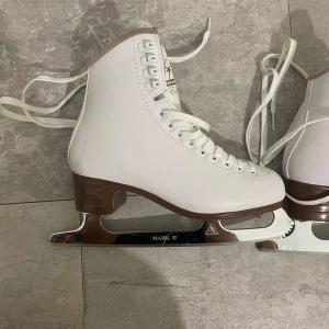 Vita konståkningsskridskor Jackson - Snygga vita konståkningsskridskor från Jackson med bruna detaljer och snörning. Modellen heter Excel och har Mark II-stål. Klassisk design med högt skaft och rund tå, perfekta för dig som vill glida fram på isen med stil.