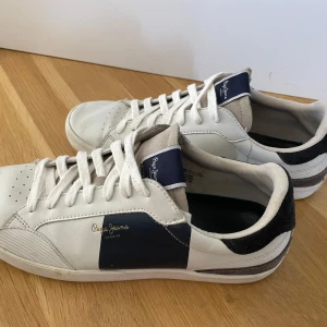 Vita sneakers från Pepe Jeans strl 42 - Snygga vita sneakers från Pepe Jeans med marinblå detaljer och svart mocka på hälen. Klassisk rund tå, snörning och diskret logga på sidan. Ovandel i skinn och textil, sula i vitt gummi. Perfekta för en clean och stilren look 😊