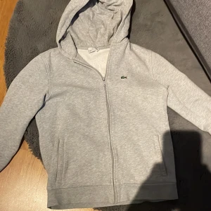 Grå hoodie från Lacoste med dragkedja - Snygg ljusgrå hoodie från Lacoste med klassisk krokodil-logga på bröstet. Tröjan har huva, hel dragkedja framtill och två fickor. Tillverkad i mjuk bomullsblandning och har långa ärmar. Perfekt för chill dagar eller när du vill ha en clean look.