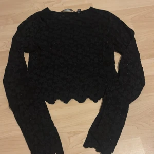 Svart spetsig croppad blus - Svart långärmad blus i spets med blommigt mönster. Toppen är croppad med vågig kant nertill och vid ärmslut, vilket ger en snygg och trendig look. Perfekt för dig som gillar detaljer och vill sticka ut lite extra.