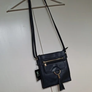 Svart axelväska från Erick Style - Snygg Mörkblå axelväska i skinnimitation från Erick Style. Väskan har guldfärgade detaljer, dragkedja framtill och baktill samt en dekorativ tofs med ring på framsidan. Justerbar axelrem och flera fack inuti gör den både praktisk och stilren.