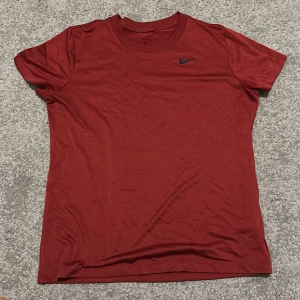 Röd Nike Dri-FIT t-shirt - Snygg röd t-shirt från Nike med klassisk svart swoosh-logga på bröstet. Tillverkad i Dri-FIT-material som andas och håller dig torr. T-shirten har rund halsringning och korta ärmar, perfekt för en sportig och avslappnad stil.