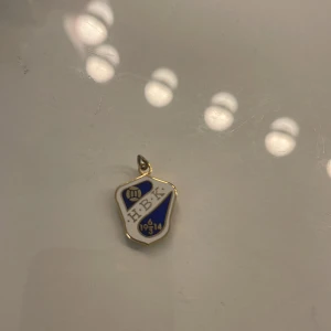 HBK halsbandshänge i blått och vitt - Unikt halsbandshänge med HBK-emblem i blått och vitt, detaljerat med guldkanter och en liten fotboll. Perfekt för dig som vill visa klubbkänsla och stil. Hänget har årtalet 1914 ingraverat och är format som en sköld.