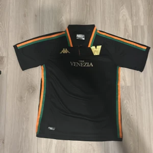 Venezia FC svart fotbollströja Kappa - Snygg svart fotbollströja från Kappa för Venezia FC med guldiga loggor och text. Tröjan har gröna och orange ränder längs axlar, krage och sidor. Klassisk krage och korta ärmar. Tillverkad i lätt och ventilerande material, perfekt för match eller träning. Använd 2 gånger 