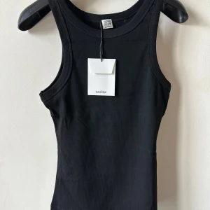 Svart ribbad linnetopp från Totême - Säljer en svart ribbad linnetopp från Totême i modellen Curved Rib Tank. Toppen har en tight passform, rund halsringning och är ärmlös. Perfekt basplagg med stilren look och diskreta sömmar. Tillverkad i mjuk bomull för maximal komfort.