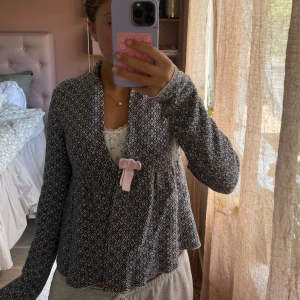 Odd Molly Mönstrad cardigan jacka med rosett - Superfin svart och vit cardigan jacka med diskret mönster och en söt ljusrosa rosett framtill. Jackan har lång ärm och en lös, flowy passform som ger en chill vibe. Perfekt att slänga över en topp för en söt och avslappnad look.