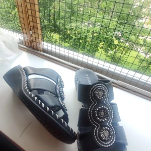 Svarta sandaler från Gyllene Gripen - Svarta sandaler med breda remmar och dekorativa broderade detaljer samt pärlliknande dekorationer framtill. Snygg kontrastsöm runt sulan och platt sula för skön känsla. Perfekta för dig som vill ha något unikt och bekvämt till sommaren.