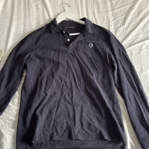 Mörkblå pikétröja från Morris - Snygg mörkblå långärmad pikétröja från Morris med klassisk krage och två knappar framtill. Tröjan har ett broderat Morris-emblem på bröstet och är gjord i mjuk bomull. Perfekt för dig som gillar stilrena och tidlösa plagg.