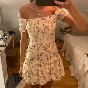 Blommig offshoulder kortklänning S - Supersöt kort klänning med offshoulder och volangkant. Klänningen är ljus med blommigt mönster i rosa, gul och grönt. Smockad överdel ger en tight passform och den har korta ärmar. 🩷🩷