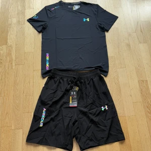 Svart Under Armour t-shirt med färgglada detaljer - Svart t-shirt från Under Armour med korta ärmar och rund hals. Loggan på bröstet och texten på sidan har en färgglad regnbågseffekt. Materialet är lätt och andas, perfekt för träning. Snygg och sportig look med coola detaljer på axel och ärm.