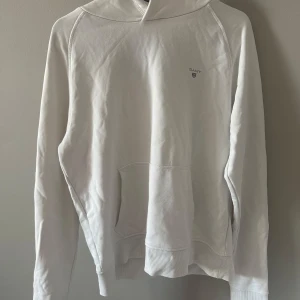 Vit hoodie från GANT med huva - Säljer en stilren vit hoodie från GANT med klassisk logga på bröstet. Tröjan har huva, magficka och långa ärmar. Perfekt för chill dagar och enkel att matcha med jeans eller joggers. Mjuk bomullskvalitet som känns skön mot huden.
