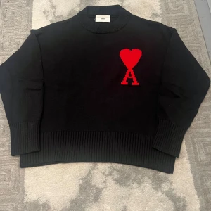 Svart stickad tröja från Ami med hjärta - Svart stickad tröja från Ami med ett stort rött hjärta och bokstaven A broderat på bröstet. Tröjan har ribbade muddar och nederkant, samt en rund hals. Perfekt för dig som gillar statement-plagg och snygga detaljer.