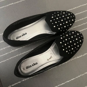 Svarta ballerinas med pärlor från Din Sko - Stilrena svarta ballerinas från Din Sko med rund tå och dekorerade med små pärlor på ovandelen. Skorna har platt sula och är perfekta för dig som gillar en klassisk look med en twist. Materialet känns mjukt och ger en elegant känsla. Använd ändast 2 gånger.
