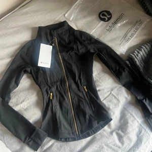 Svart Define jacka från Lululemon - Snygg svart träningsjacka från Lululemon med guldiga dragkedjor och figurnära passform. Jackan har hög krage, långa ärmar och två fickor med dragkedja. Jackan är oanvänd och lappen sitter kvar i jackan. Lululemon dustbag följer med.