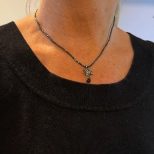 Svart glittrigt halsband med blomma - Snyggt svart halsband med pärlor och en blomformad silverfärgad hängare med stenar. Hängaren har en rund svart pärla som detalj. Perfekt för dig som gillar unika smycken med lite bling och cool design.