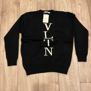 Svart stickad tröja från Valentino - Svart stickad tröja från Valentino med vit VLTN-logga i stort tryck längs framsidan. Klassisk rund halsringning och långa ärmar. Perfekt för dig som gillar stilrena designer med en lyxig touch.