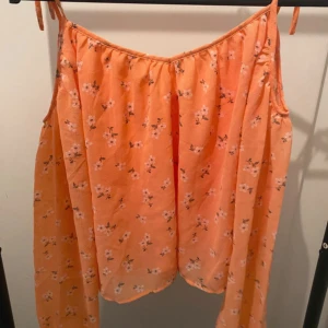 Blommig orange linnetopp från H&M Divided - Superfin orange linnetopp med små vita blommor från H&M Divided. Toppen har tunna axelband med knyt och en loose passform som ger en chill vibe. Perfekt för varma dagar och sommarhäng. Materialet är lätt och känns skönt mot huden.