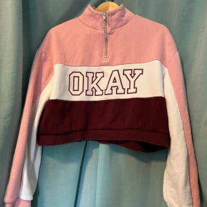 Croppad rosa och vinröd tröja med zip - Snygg croppad tröja i rosa, vitt och vinrött med texten 'OKAY' framtill. Tröjan har lång ärm, half zip med ring och hög krage. Perfekt för dig som gillar sportig och trendig stil.