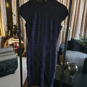 Mörkblå spetsklänning med omlott - detalj - Supersnygg mörkblå klänning i spets med blommigt mönster och omlott-detalj framtill. Klänningen har korta ärmar och är vadlång, perfekt för dig som vill ha en elegant look. Materialet är mjukt och följsamt, och spetsen ger en lyxig känsla.