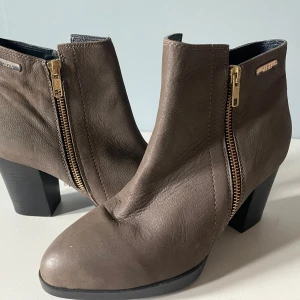 Bruna stövletter med klack från Dasia - Snygga bruna stövletter från Dasia i äkta skinn med guldfärgade dragkedjor på sidorna. Skorna har en stabil klack och rund tå, perfekt för dig som vill ha en stilren och trendig look. Klassisk modell som funkar till många outfits.