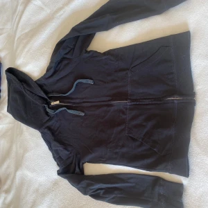 Svart hoodie med dragkedja och huva - Svart hoodie med huva och snörning, hel dragkedja framtill och två fickor. Mjuk bomullskänsla och ribbade muddar vid ärmslut och nederkant. Perfekt för chill dagar eller när du vill ha något bekvämt och enkelt.