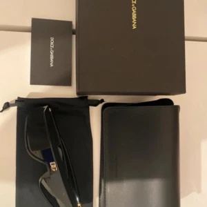 Svarta solglasögon Dolce & Gabbana - Snygga svarta solglasögon från Dolce & Gabbana med bred, futuristisk båge och DG-logga i guld på sidan. Kommer med originalfodral, extra lins och förvaringspåse. Perfekta för dig som vill sticka ut med en lyxig accessoar.