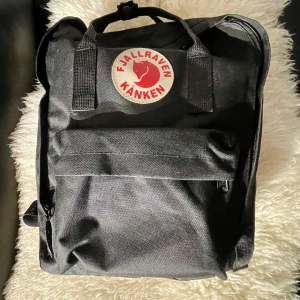 Svart Fjällräven Kånken ryggsäck - Klassisk svart Fjällräven Kånken ryggsäck med ikonisk rund logotyp framtill. Rymligt huvudfack, mobil, flaska fack , framficka med dragkedja och två sidofickor. Tillverkad i slitstark syntet med justerbara axelremmar och topphandtag. Perfekt för skola, city eller utflykter.