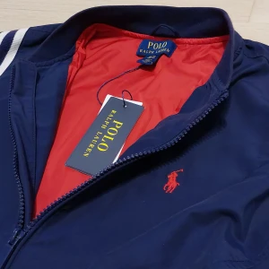 Marinblå vindjacka Polo Ralph Lauren - Snygg marinblå vindjacka från Polo Ralph Lauren med röd insida och klassisk logga broderad på bröstet. Jackan har vita ränder på axlarna, dragkedja framtill och två fickor med dragkedja. Perfekt för dig som gillar sportig och stilren look.