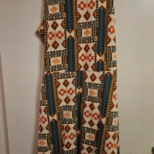 Mönstrad klänning från H&M - Färgstark klänning från H&M med geometriskt mönster i orange, rött, mörkgrönt och beige. Klänningen är ärmlös och har en luftig passform, perfekt för dig som gillar unika prints och bohemisk stil. Materialet känns lätt och bekvämt mot huden.