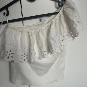 Vit offshoulder topp med volang Zara - Superfin vit offshoulder topp från Zara med bred volangkant och broderade blommiga detaljer. Toppen är ärmlös och har en romantisk känsla med snyggt hålmönster längs volangen. Perfekt för sommarens alla tillfällen!