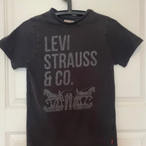 Svart Levi's t-shirt med tryck - Cool svart t-shirt från Levi's med stort grått tryck framtill där det står 'Levi Strauss & Co.' och en grafisk illustration med hästar. Klassisk rund hals och korta ärmar. Mjuk bomullskvalitet och röd Levi's-tag på sidan. Perfekt till jeans och sneakers.