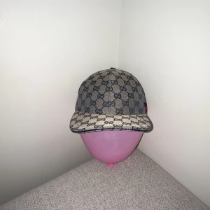 Gucci keps med GG-mönster och röd-blå rand - Snygg keps från Gucci i beige med klassiskt GG-monogram över hela kepsen. Kepsen har en böjd skärm och en ikonisk röd-blå rand på sidan. Tillverkad i bomullsmix med justerbar rem baktill. Perfekt för dig som vill sticka ut med en lyxig accessoar.