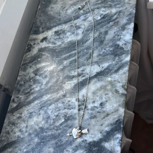 Silverhalsband med hjärta och pil - Snyggt halsband i silverfärg med ett hjärtformat hänge som genomborras av en pil. Pilens fjädrar har små glittriga detaljer som ger extra bling. Perfekt för dig som gillar romantiska och trendiga smycken med lite edge. Halsbandet är Nickelfritt. Aldrig använt.