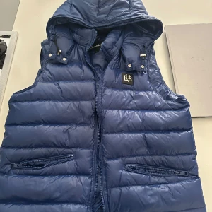  BVALDI väst  - Snygg blå dunväst från BVALDI med quiltad design och avtagbar huva. Västen har två dragkedjefickor framtill och en svart logotyp-patch på bröstet. Perfekt för lager-på-lager och chill stil under höst och vinter.den är helt ny om du är intresserad är det bara att  skriva