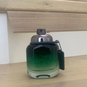Coach herrparfym grön glasflaska - Coach herrparfym i en snygg grön glasflaska med metallfärgat spraylock och Coach-logga ingraverad på framsidan. Flaskan har en cool gradient från mörkgrönt till klart glas och en svart tag i kedja hänger på sidan. Perfekt för dig som gillar stilrena och moderna dofter.