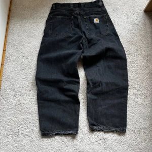 Carhartt Brandon pants  - Säljer ett par svarta jeans från Carhartt med bred passform och raka ben. Byxorna har två stora bakfickor och en med Carhartt-logga, och råa, lite slitna benslut. Perfekta för dig som gillar loose och avslappnad stil.