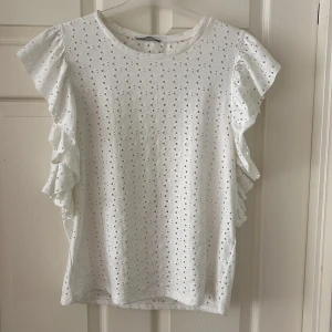 Vit topp med volangärmar, Zara - Säljer en söt vit topp med broderade hålmönster och volangdetaljer längs ärmarna. Blusen har rund hals och är tillverkad i mjuk bomull, perfekt för en fräsch och somrig look. Passar dig som gillar romantiska detaljer och ljusa färger. Använd typ 4 gånger. 