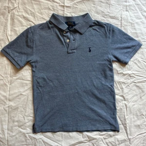 Blå Polo Ralph Lauren pikétröja - Klassisk blå pikétröja från Polo Ralph Lauren barnstorlek M (10-12). Tillverkad av mjuk och slitstark bomull med klassiska detaljer: • klassisk Polo Ralph Lauren broderad logotyp på bröstet • Klassisk krage med knappslå  📏 Mått (plattliggande): • Längd: ca 54 cm • Bröstvidd: ca 40 cm  Skick: Mycket bra – inga fläckar eller hål. Perfekt för både vardagsbruk och smarta casual-tillfällen.