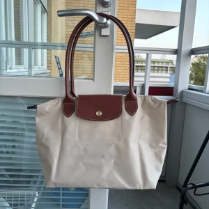 Beige axelväska från Longchamp - Snygg beige axelväska från Longchamp med bruna skinnhandtag och lock. Väskan har guldfärgad dragkedja med Longchamp-logga och klassisk stilren design. Perfekt storlek för vardagens alla grejer och enkel att matcha med olika outfits.