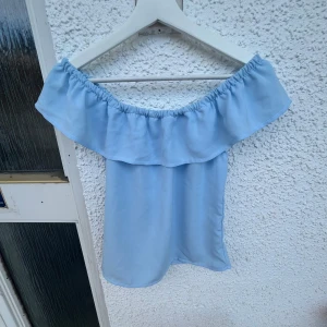 Ljusblå offshoulder volangtop - Supersöt ljusblå offshoulder topp med bred volang runt hela axelpartiet. Tunn och luftig, perfekt för varma dagar. Sitter snyggt tack vare resår upptill. Enkel att matcha med både jeans och kjol för en fräsch look.